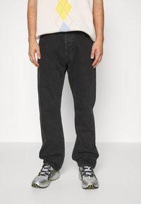 Osoba w czarnych, luźnych dżinsach, białych trampkach z czarnymi i żółtymi akcentami oraz w kremowym swetrze w żółto-niebieski wzór w romby.