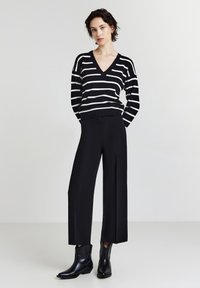 Donna in piedi con le mani dietro la schiena, con indosso un maglione con scollo a V a righe bianche e nere, pantaloni larghi neri e stivaletti neri.