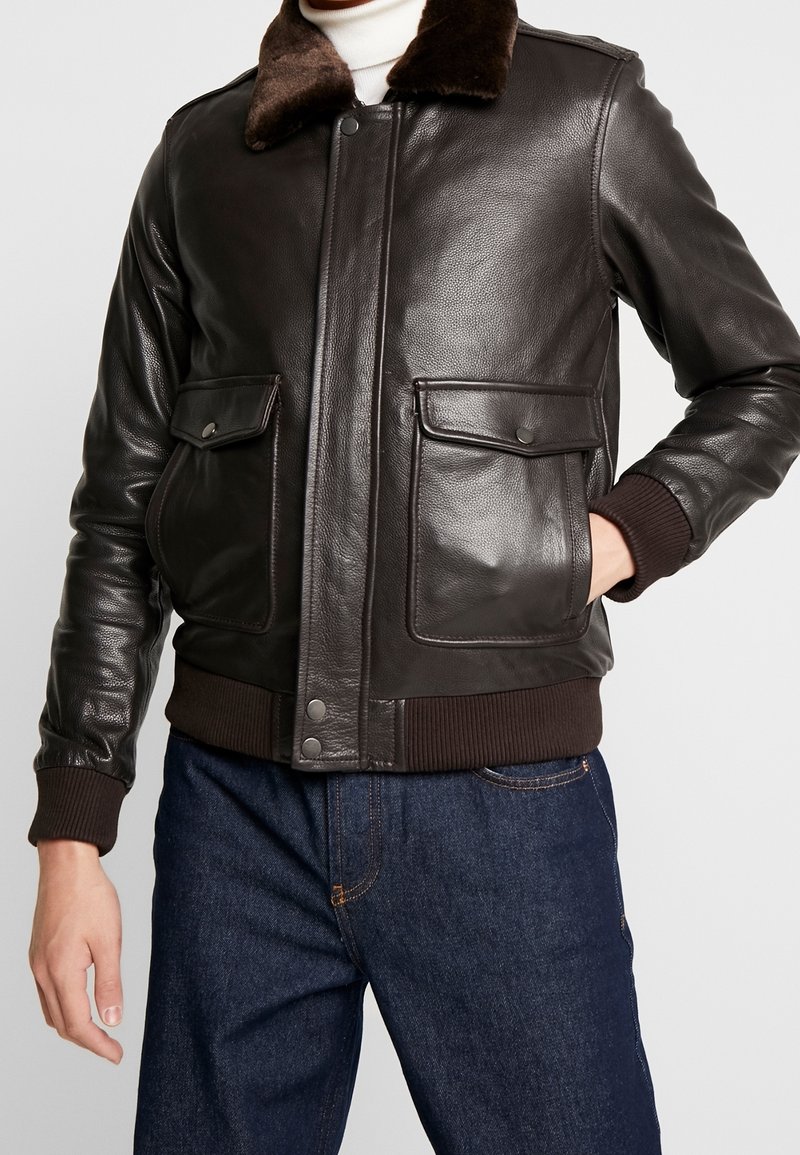 Hombre con chaqueta tipo bomber de cuero marrón oscuro con cuello de piel y bolsillos frontales con solapa, combinado con vaqueros azules y un suéter de cuello alto blanco.