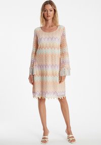 Robe multicolore jusqu'au genou avec un motif en zigzag dans des teintes pastel. Dotée de manches longues avec des poignets à franges et d'un col rond.
