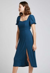 Robe teal avec décolleté carrée et manches bouffantes. Fabriquée en tissu texturé, elle présente une fente latérale et une taille cintrée, arrivant au milieu du mollet.
