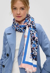 Écharpe multicolore présentant un motif floral avec des accents bleu, rose et blanc. Tissu léger avec des bords effilochés, enroulée autour du cou d'un modèle.