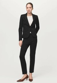 Kobieta stojąca w czarnej, dopasowanej marynarce z wąskimi spodniami, białym topem i brązowymi, niskimi butami na obcasie o szpiczastym nosku na jednolitym jasnym tle.