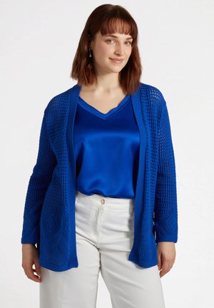 Cardigan blu lavorato a maglia con motivi a rombi e design a trama aperta. Indossato sopra una blusa blu in satin a scollo a V, abbinato a pantaloni bianchi.