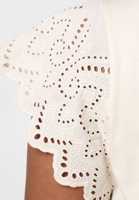 Blouse en ivoire avec des manches en dentelle brodée délicate à bords festonnés et motifs découpés, texture de tissu douce.