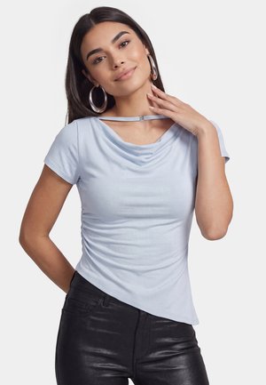 Junge Frau mit langen dunklen Haaren, die große Creolen trägt, ein hellblaues Kurzarm-Top mit dekorativem Ausschnitt und schwarze Hose.