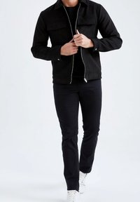 Giacca nera in suede con due tasche sul petto, chiusura con zip e maniche lunghe, abbinata a pantaloni neri slim fit e sneakers bianche.