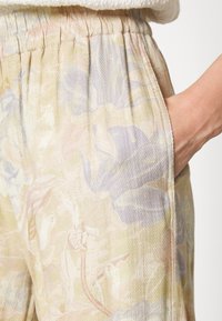 Gros plan d'une main dans la poche d'un pantalon beige à motif floral texturé, avec une taille élastique et des couleurs pastel douces.