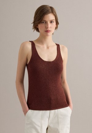 Vrouw met kort bruin haar draagt een mouwloos bruin gebreid topje en een witte broek, staand met handen in de zakken tegen een neutrale achtergrond.