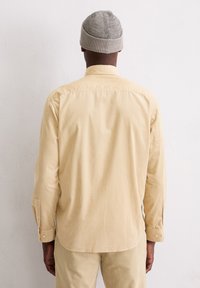 Chemise en velours côtelé beige clair à manches longues, avec des poignets à boutons et un col classique, portée avec un pantalon assorti et un bonnet en côte gris.