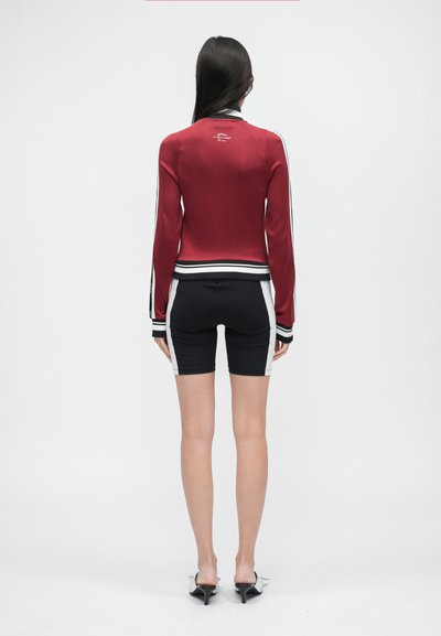 Giacca bomber in raso rosso con dettagli a righe nere e bianche, abbinata a pantaloncini neri a vita alta con pannelli laterali bianchi e design aderente.