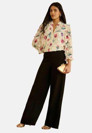 Kvinde iført en tynd blomstret bluse med voluminøse ærmer, sorte bukser med vide ben, gyldne sandaler, gyldne øreringe og holder en gylden clutch.