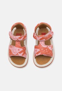 Vaaleanpunaiset sandaalit, joissa on kissan ja kalojen kuvioita, varustettu tarranauhalla ja pehmustetuilla pohjilla, asetettu valkoiselle pohjalle, jossa on oransseja yksityiskohtia.