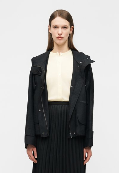 Femme portant une veste à capuche noire sur un haut crème boutonné et une jupe plissée noire, faisant face à l'avant sur un fond uni.