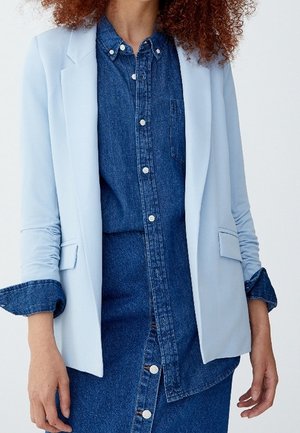 Kvinde iført en lyseblå blazer over en mørkeblå denim skjorte med knapper og matchende denim nederdel, stående mod en ensfarvet baggrund.
