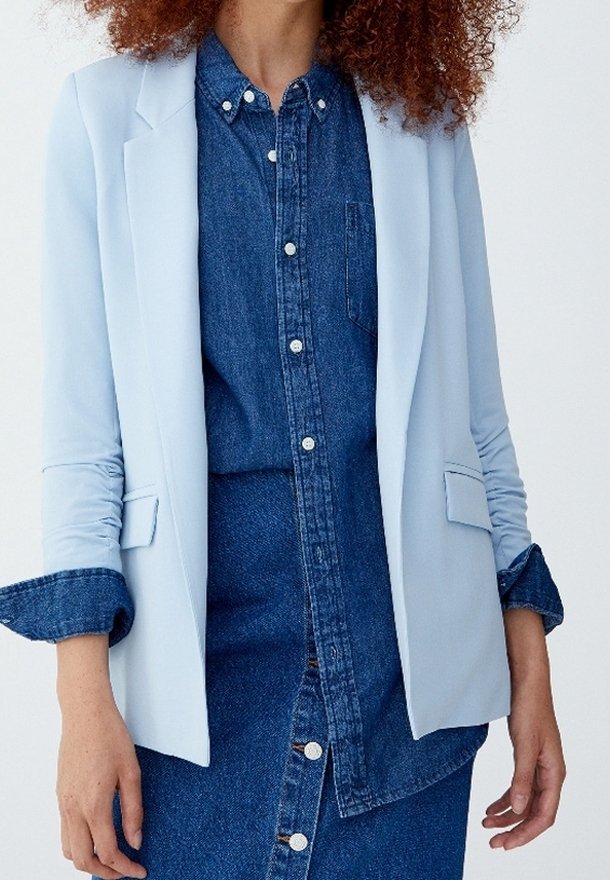 Femme portant un blazer bleu clair par-dessus une chemise en jean bleu foncé boutonnée et une jupe en jean assortie, debout devant un fond uni.