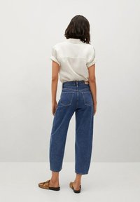 Camisa ligera de crema con mangas cortas, combinada con unos vaqueros azules de talle alto y holgados, y sandalias marrones de slide. Textura suave con mangas enrolladas.