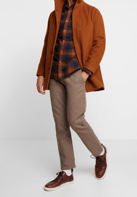 Abrigo de lana color óxido, camisa a cuadros en azul marino y naranja, pantalones beige y zapatillas de cuero marrón con suelas blancas.