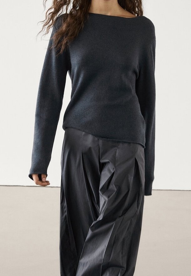 Femme portant un pull en maille à manches longues gris foncé et un pantalon noir large à taille haute, ample, sur un fond uni.