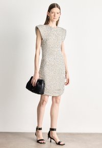 Vestido prateado cintilante com lantejoulas, mangas em capa, design justo ao corpo, superfície texturizada, combinado com uma mala preta plissada e saltos com tira ao tornozelo.