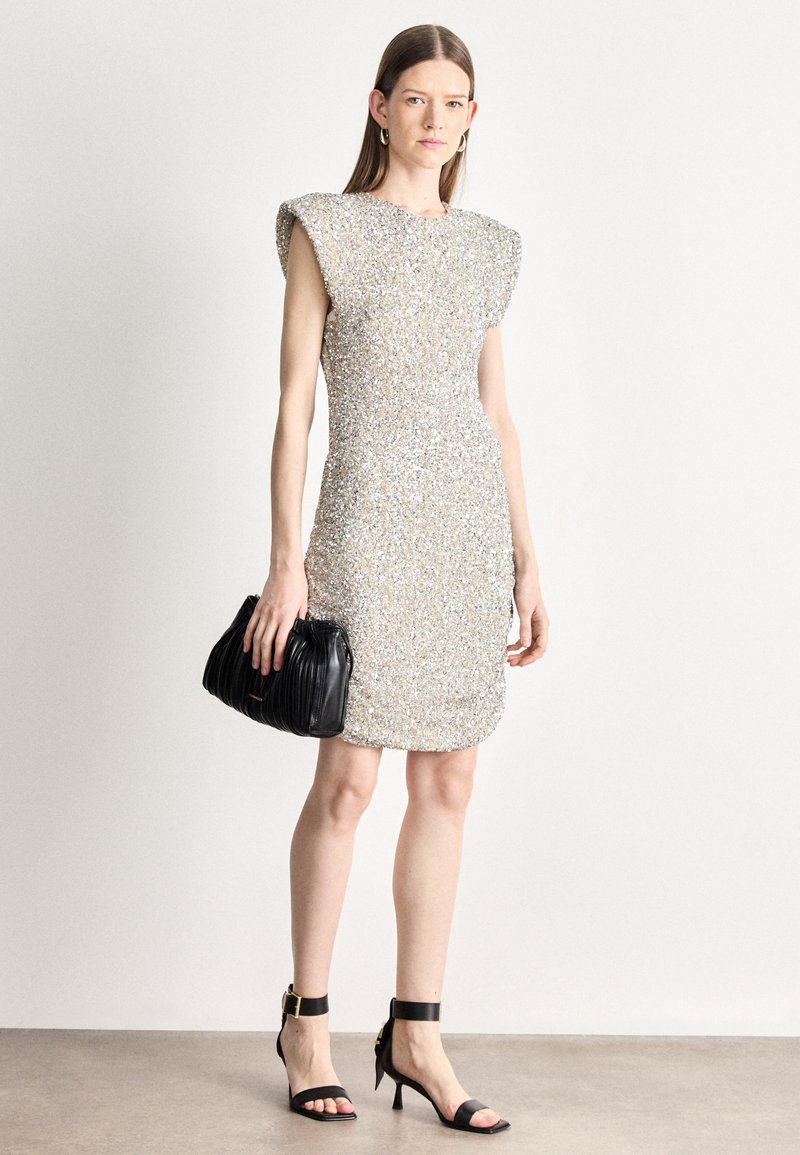 Vestido prateado cintilante com lantejoulas, mangas em capa, design justo ao corpo, superfície texturizada, combinado com uma mala preta plissada e saltos com tira ao tornozelo.