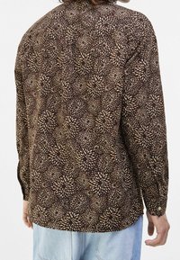 Brun mönstrad skjorta med blommönster och ljusa accenter; långa ärmar med knappmanchetter; avslappnad passform; tygtextur synlig.