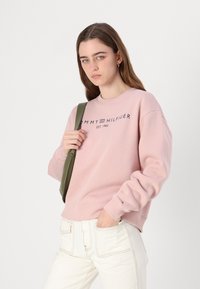 Růžová mikina s kulatým výstřihem a dlouhými rukávy, zdobená tmavě modrým nápisem "TOMMY HILFIGER" a logem, nošena s světlými džíny a zelenou taškou přes rameno.