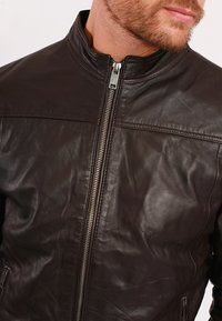 Brun läderjacka med hög krage, framsidans dragkedja och två sidofickor. Texturerad yta med lätt glans och rena sömnadsdetaljer.