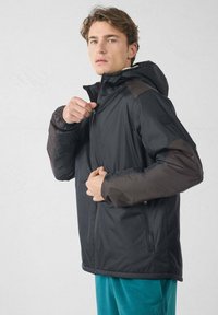 Jonge man met lichtbruin haar die een zwart getailleerd jack met bruine patches op de mouwen dichtritst, en een blauwe broek draagt.