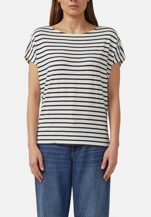 Femme portant une chemise blanche à rayures horizontales noires et un jean bleu, debout devant un fond clair uni.