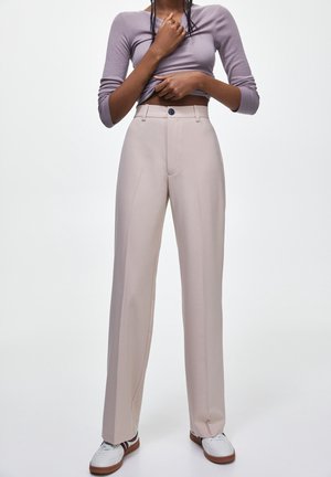 Persoon die beige hoge taille broek met wijde pijpen draagt, een lavendelkleurige top met lange mouwen omhoog tot de buik, en witte sneakers met bruine zolen.