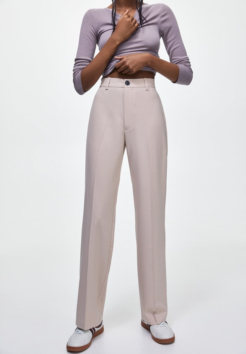 Personne portant un pantalon beige taille haute à jambes larges, un haut à manches longues lavande relevé sur le ventre, et des baskets blanches avec des semelles marron.