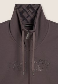 Giacca grigia con zip e colletto alto. Presenta dettagli metallici con la scritta "FRE DDY" e un motivo interno testurizzato.