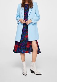 Donna che indossa un blazer azzurro chiaro sopra un vestito floreale scuro con uno spacco alto fino alla coscia, abbinato a stivali bianchi alla caviglia con tacco a blocco.