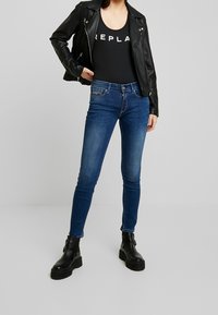 Jeans skinny de color azul oscuro con una cintura de altura media, que presentan un ligero efecto de desgaste y un estilo clásico de bolsillos. Combinados con una camiseta sin mangas negra y una chaqueta de cuero.