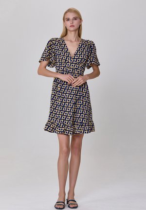 Robe marine avec un motif géométrique doré, manches courtes bouffantes, décolleté en V, taille cintrée et ourlet froncé. Portée avec des sandales plates.