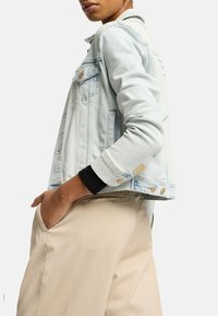 Lyseblå denimjakke med en tætsiddende pasform, udstyret med guldhardware, to brystlommer og en kontrastfarvet sort ærme nedenunder.