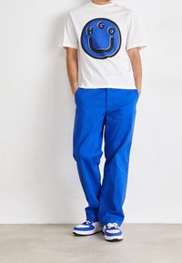 Camiseta blanca con un gráfico circular en azul y negro que presenta las letras "HGO". Combinada con pantalones rectos de un azul brillante y zapatillas con acentos en azul.