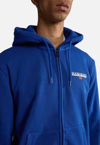 Blå zip-up hættejakke med snoretræk i hætten, med et logo-patch på brystet, lavet af tekstureret stof og har frontlommer.