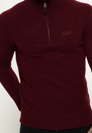 Mand iført en tætsiddende vinrød strikket pullover med kort lynlås foran og høj hals.