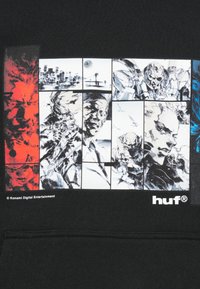 Melns džemperis ar kapuci, uz kura ir grafiskais attēls ar vairākiem komiksiem līdzīgiem ilustrācijām melnbaltā krāsā, papildināts ar sarkaniem un ziliem akcentiem.