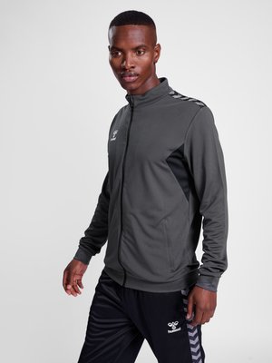 Mann trägt eine graue und schwarze Sportjacke mit Reißverschluss und schwarze Sporthose mit Chevronstreifen, steht vor schlichtem Hintergrund.