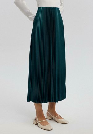 Femme portant une jupe midi plissée vert foncé, un haut blanc à manches longues et des chaussures beige à talons bas avec brides, debout devant un fond uni.