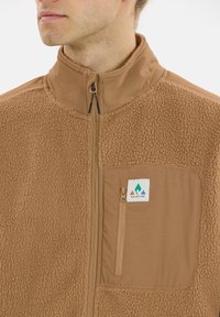 Veste en polaire marron avec un col haut, comportant une poche poitrine zippée et un écusson logo. Surface texturée et détails de panneaux lisses.
