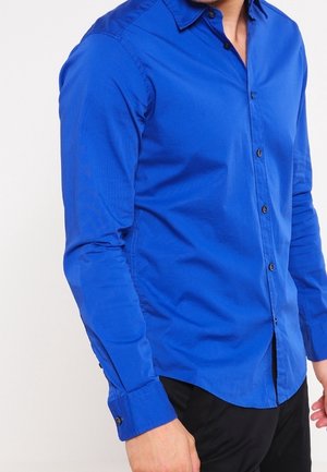 Homme portant une chemise bleue ajustée à manches longues avec un pantalon noir, debout devant un fond clair uni.