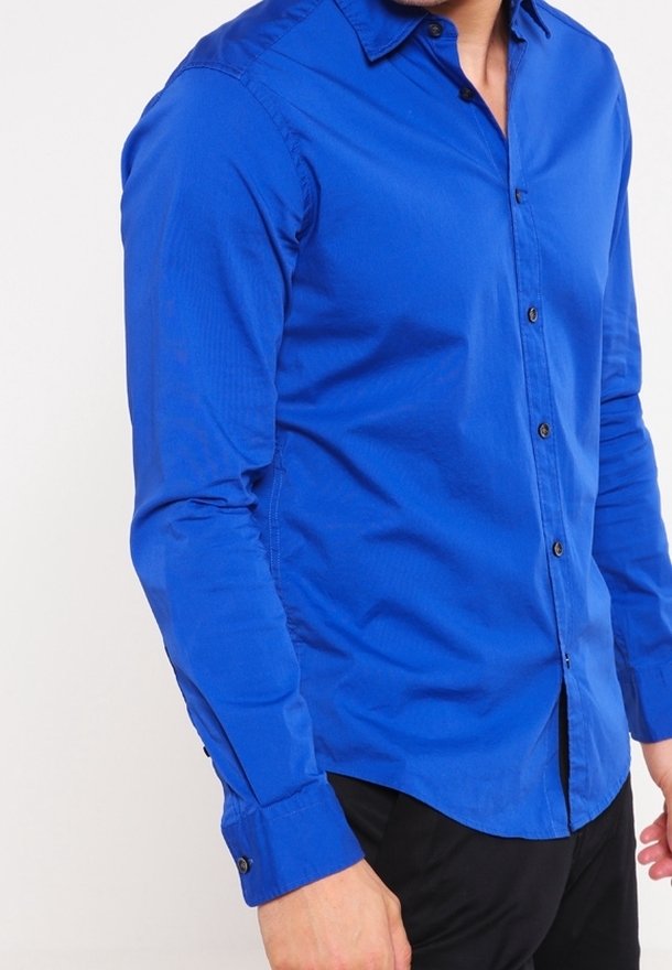 Homme portant une chemise bleue ajustée à manches longues avec un pantalon noir, debout devant un fond clair uni.
