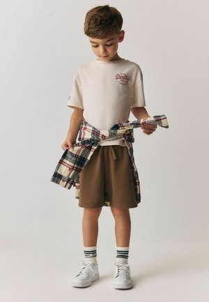 Noor poiss, kandes beeži t-särki, pruuneid lühikesi pükse ja valgeid vabaajajalatsid, seisab ja kohandab vöökohale seotud ruudulist särki ühevärvilise tausta ees.