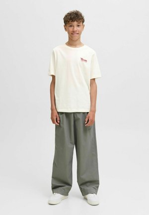 Locker sitzendes, hellbeiges T-Shirt mit rotem Logo, kombiniert mit übergroßen grauen Hosen und weißen Sneakers. Lässiges, entspanntes Outfit.