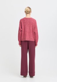 Maglione rosa lavorato a maglia con spalle scese e pantaloni bordeaux larghi con gambe ampie e orli arrotolati, visti da dietro.