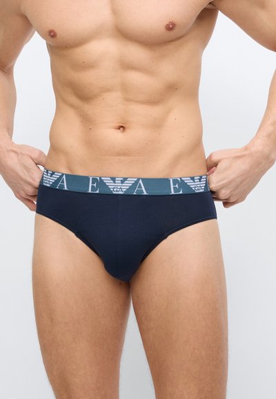 Ανδρικός κορμός με μπλε σκούρα εσώρουχα με ελαστική μέση με λογότυπο, χέρια που ρυθμίζουν τη μέση, ανοιχτόχρωμος τόνος δέρματος, μυώδης σωματού.